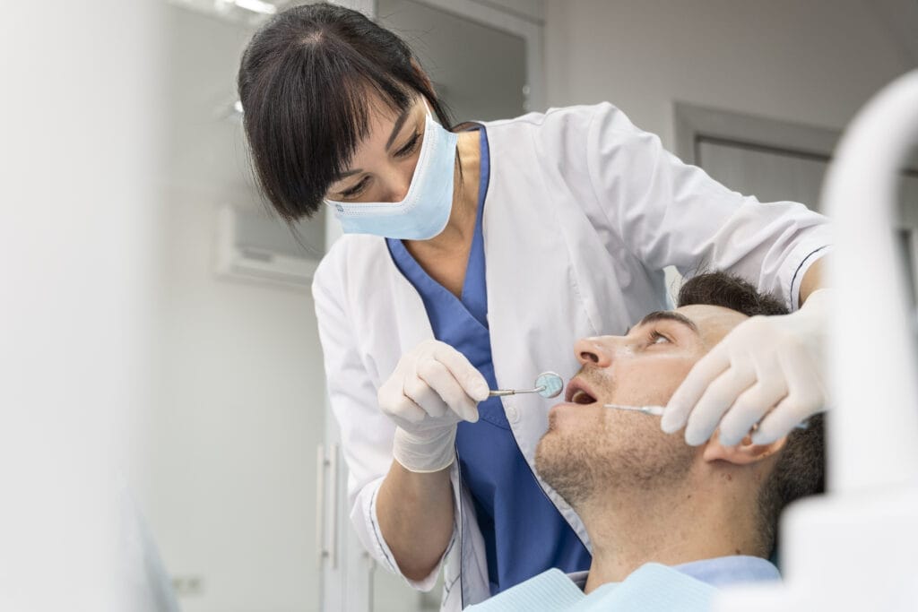 A dentist wearing a white coat and mask examines a male patient using dental tools in a bright, modern clinic.