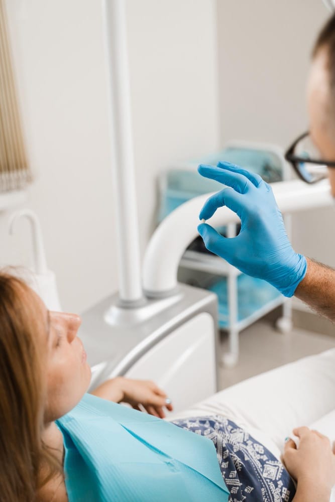 A dentist explaining a small dental item to a female patient seated in the dental chair.