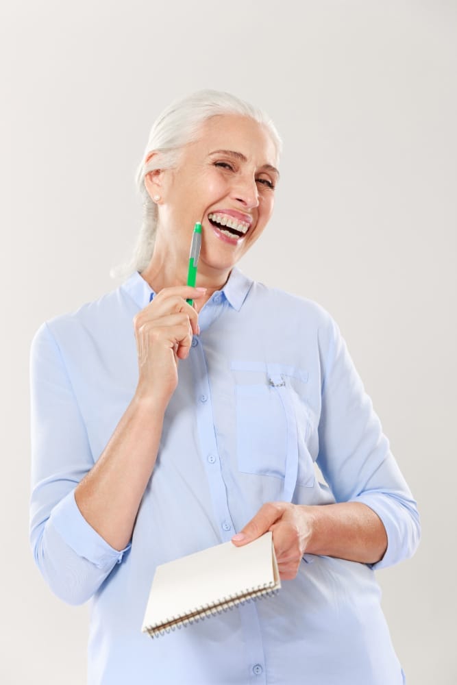 An older woman smiling and holding a notepad with a pen in hand